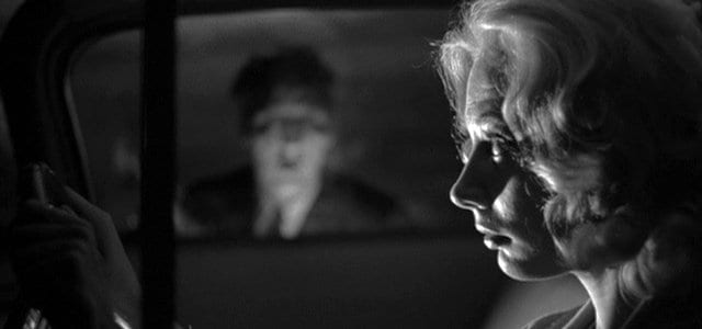 a blonde female drives her car, a blurred male face peers into her window