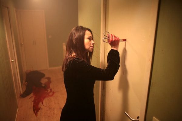 The Woman stabs bloody scissors into the door with a bloody corpse on the floor in the corridoor