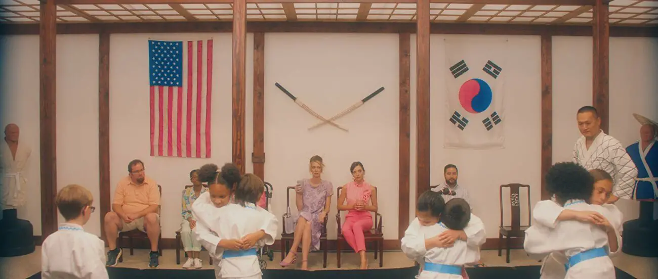 Lisa (left) and Jill (right) sit next to each other while their are in a karate class. There are children hugging in the foreground.