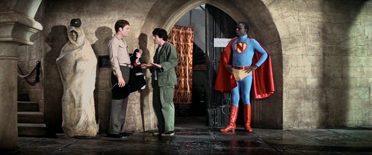 (L-R) Cl. Kane (Stacey Keach) hands Lt. Reno (Jason Miller) his SS costume as Maj. Nammack (Moses Gunn) struts in his new Superman-esque costume. 