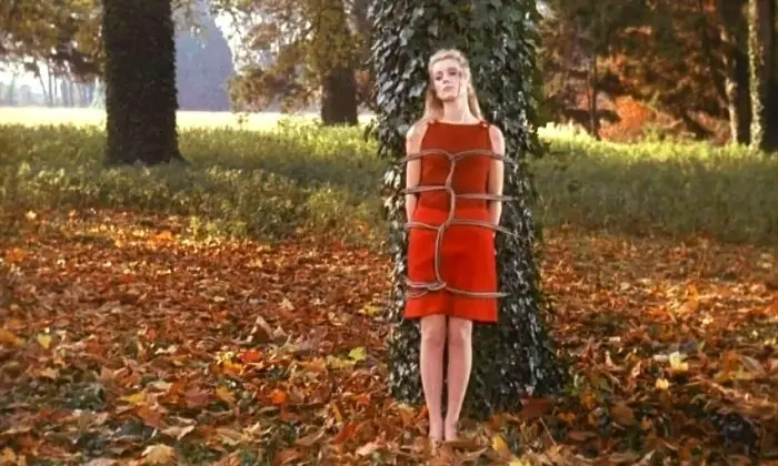 Séverine in a red dress is tied to a tree outside during one of her sexual fantasies
