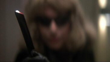 Man disguised as a woman in a blonde wig and black sunglasses holds out a razor menacingly.