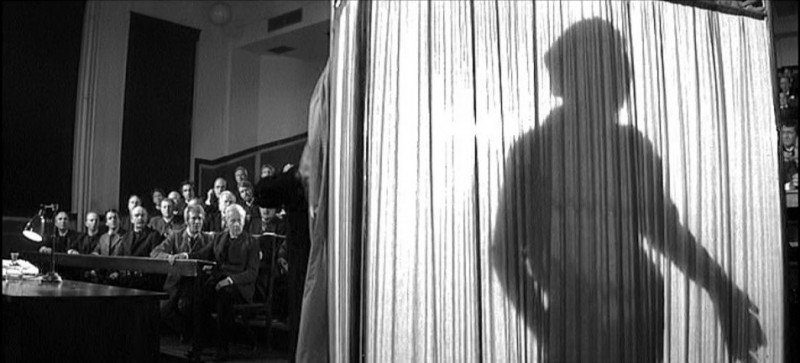 An audience looks on as the silhouette of John Merrick shows him standing behind a curtain