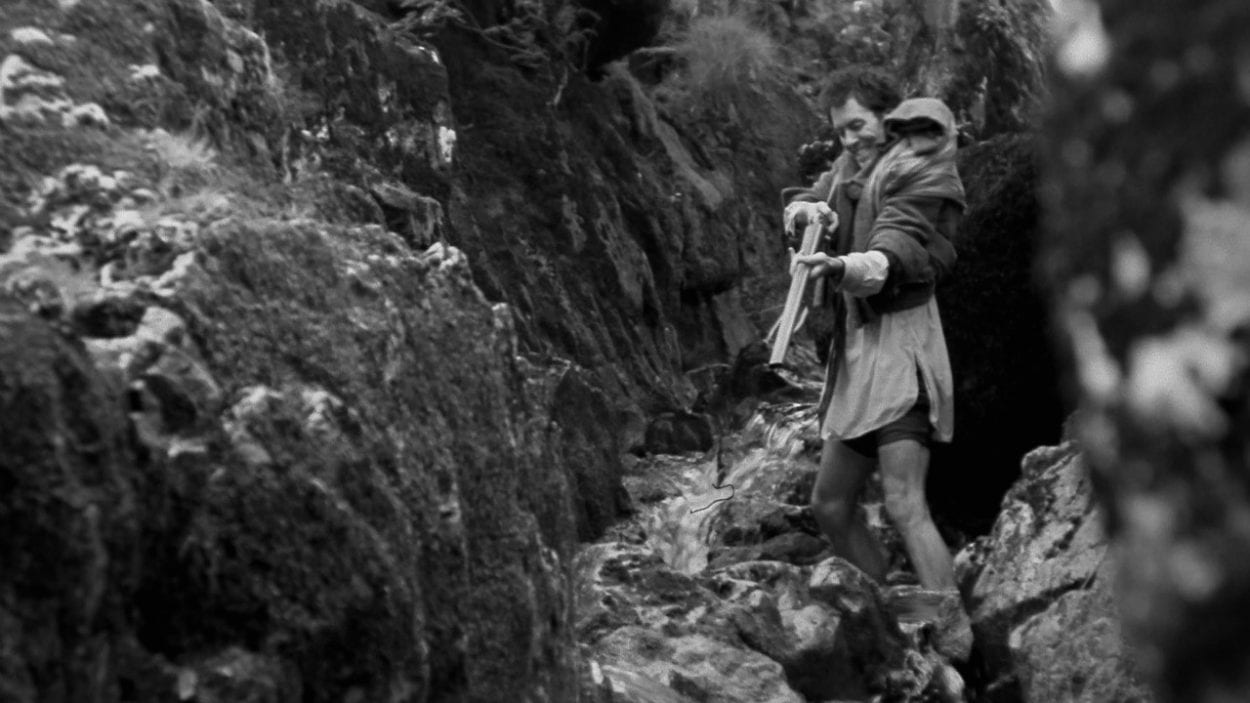 Withnail in feral mode trying to catch a fish by shooting it with a shot gun, standing in a stream
