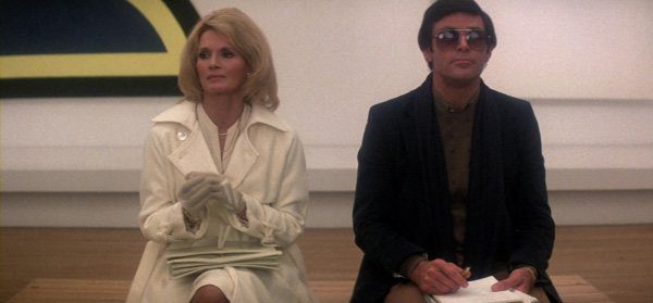 Blonde woman in white suit and man in dark suit and sunglasses sit next to each other in an art museum.
