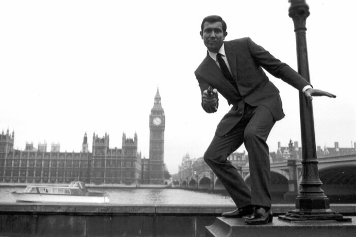 George Lazenby posing on a wall as Bond, with The Houses of Parliament in the background