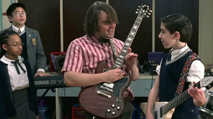 Dewey Finn (Jack Black) plays an electric guitar beside his student Zack. Behind Dewey are his students Lawrence and Alicia.