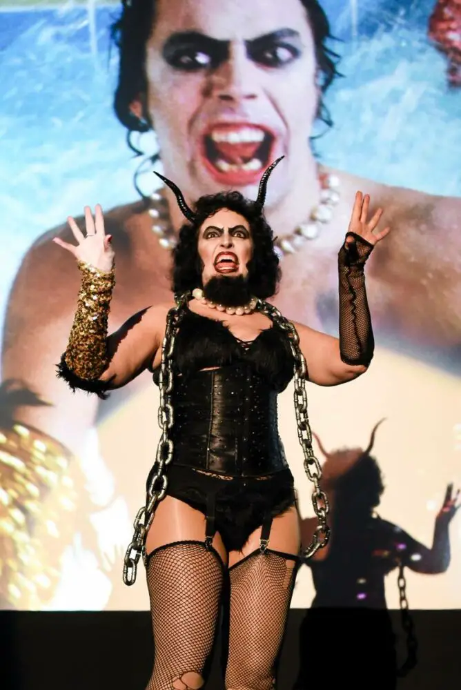 Frank-N-Furter with Krampus horns, beard and chain, singing and dancing in front of movie screen.