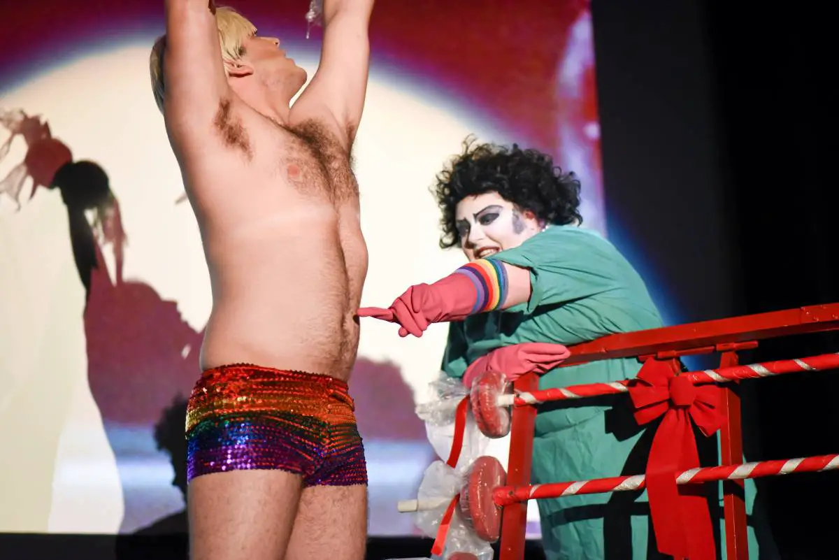 Frank-N-Furter touches Rocky's stomach, while Rocky lifts weights wearing sparkly rainbow shorts.