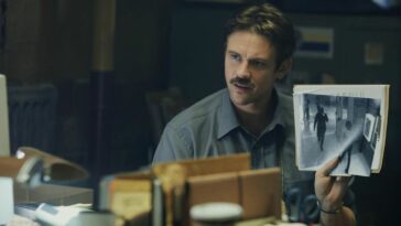 An obsessed detective holds up a still photograph at his desk