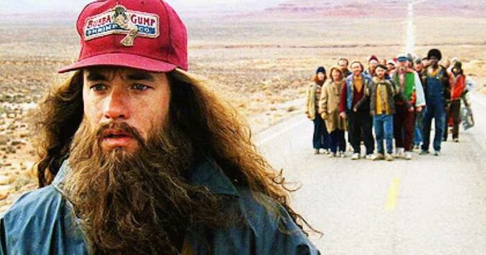 Forrest, with long hair and beard walks down a country road with a crowd following him. 