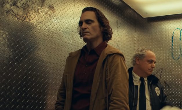 Joaquin Phoenix as Fleck, stands in an elevator with his eyes closed and a cop stands behind him