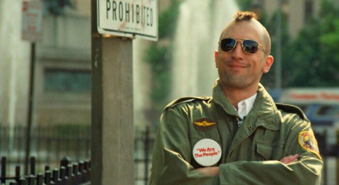 A mohawked Travis Bickle in army clothes stands and smirks while watch candidate Charles Palantine speak
