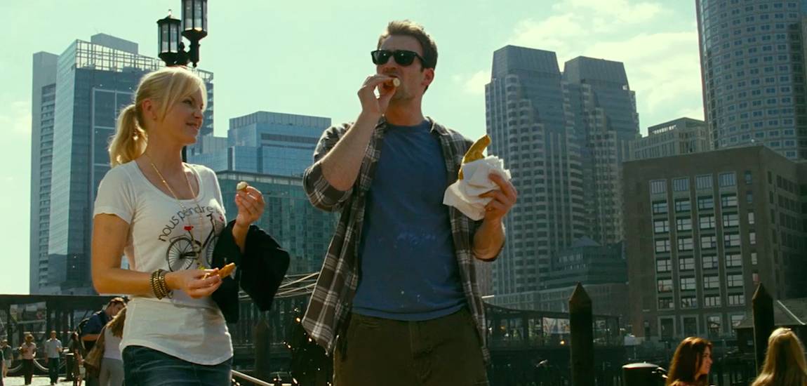 Colin eats a pretzel while walking with Ally through a park with the city of Boston in the background