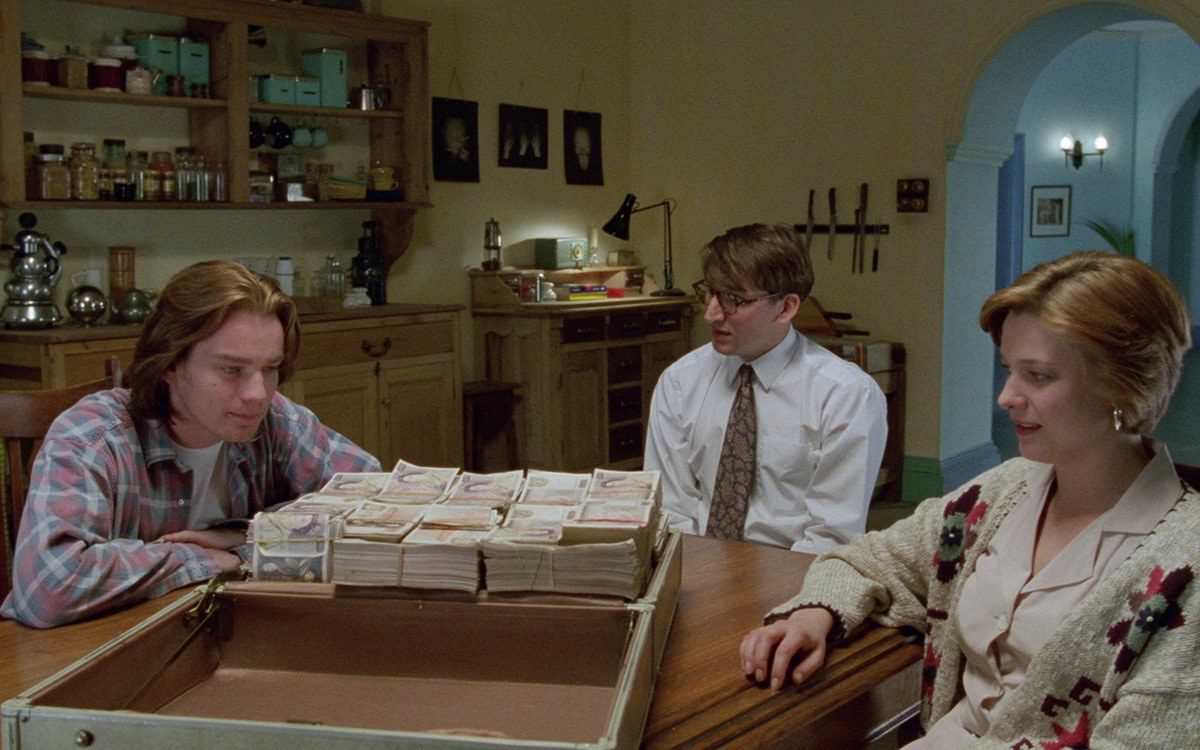 Ewan McGregor, Christopher Eccleston, and Kerry Fox sit at a table with a briefcase full of money