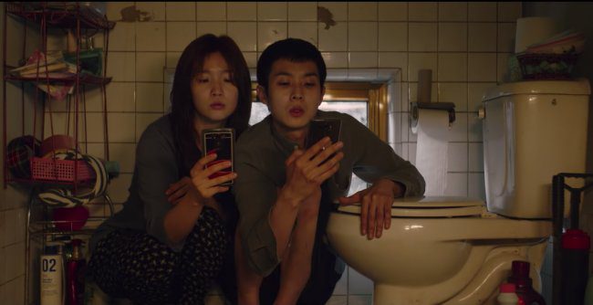 A boy and girl look at their phones as they sit by a toilet