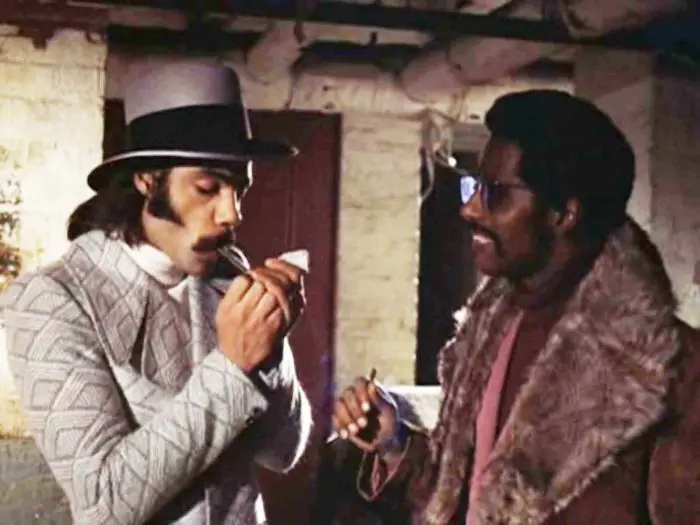Priest (Ron O'Neal) and Eddie ( Carl Lee) are stood in an alleyway having a conversation. Priest is lighting a cigar as Eddie looks on, smiling, in Superfly