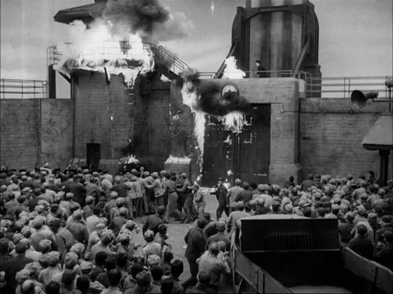 The prison tower is in flames in the background, with a crowd of inmates in foreground