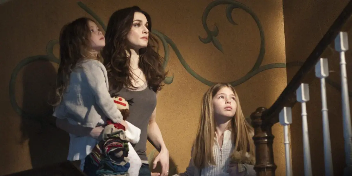 Rachel Weisz with her onscreen daughters on a staircase, yellow wall with a twisting vine painted on, all three girls with sad but accepting gazes as the girls look up and Weisz stares straight ahead