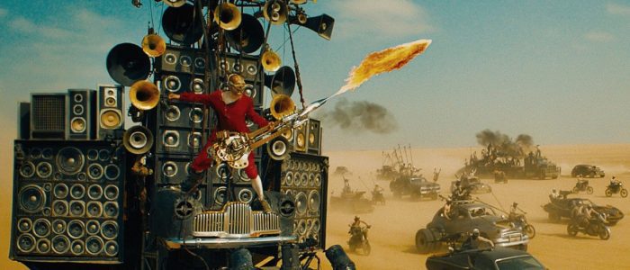 The Doof Warrior plays his fire guitar surrounded by lots of cars in the desert of Fury Road