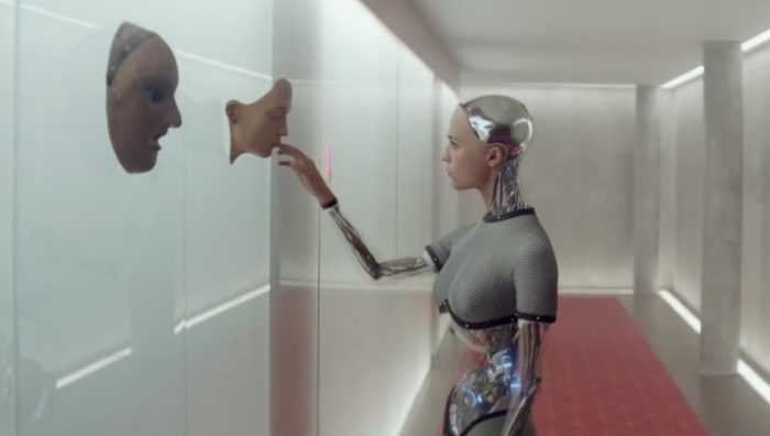Ava touches a copy of her face on a shiny wall in a bright hallway