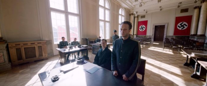 Franz stands at a table with his lawyer surrounded by Nazi insignia