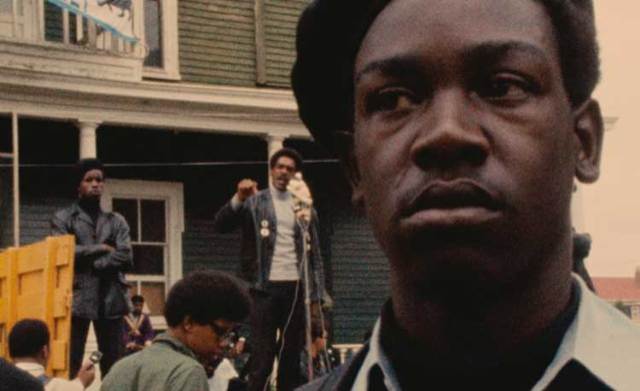 A Black Panther stands guard as a man speaks in the background to a crowd