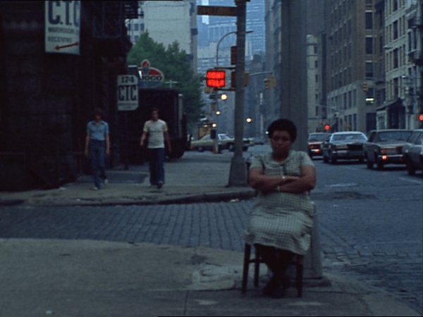 A woman sits with her hands folded on a street corner with two men walking in the background