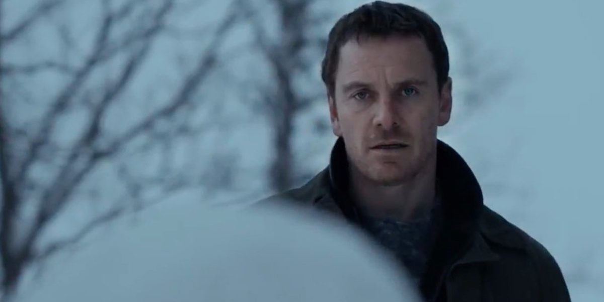 Michael Fassbender In The Snowman looking at the snowman in the forefront of the picture, its back to the viewer with trees behind Fassbender and a white cloudy background