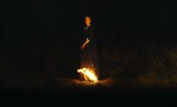 a woman stands in the dark with the tail of her dress on fire