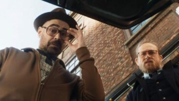 Two men with glasses look down into the trunk of a car