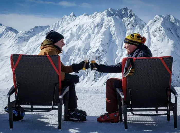 Zach and Pete sit in front of a mountain, clinking glasses of beer together