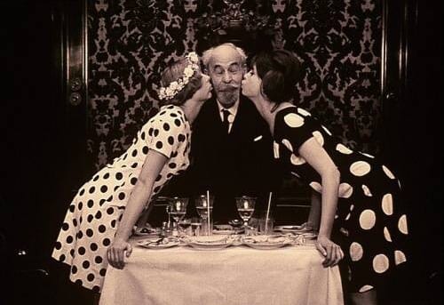 Marie I and Marie II each kiss a cheek of an old man in parallel polka dot dresses