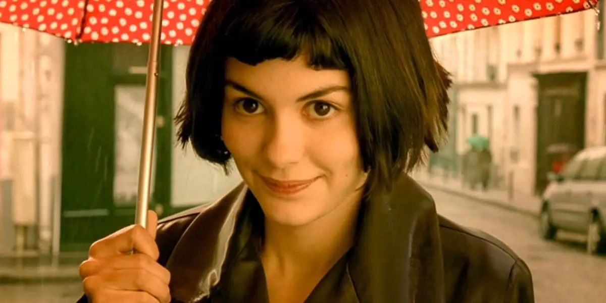 Amelie staring ahead, with smile on her face, rain in background, holding a red umbrella with white polka dots on it
