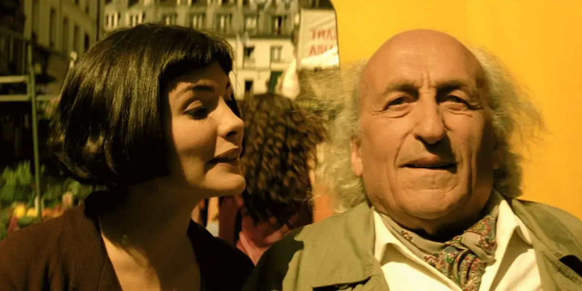 Amelie looking at the man and talking to him, the man staring straight ahead in wonder, yellow building behind them and a woman with her back to them in the background between Amelie and the man