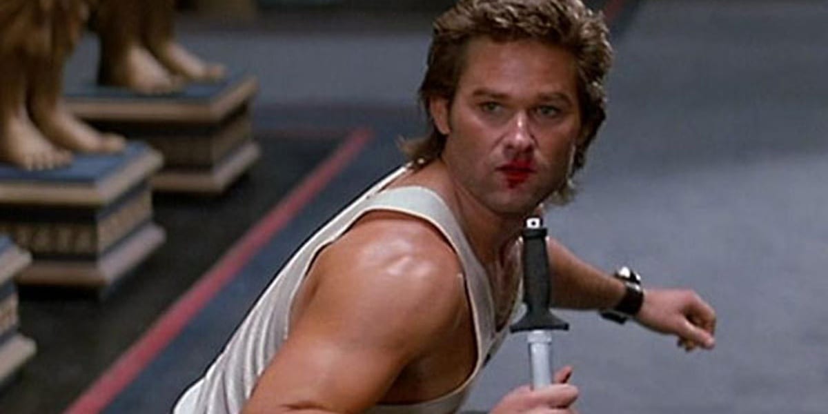Kurt Russell as Jack Burton in Big Trouble in Little China looking behind him with lipstick on his face and a knife pointing downward in his hand