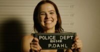 Zoey Dutch as Peg holds up a police sign and grins while getting her mugshot taken