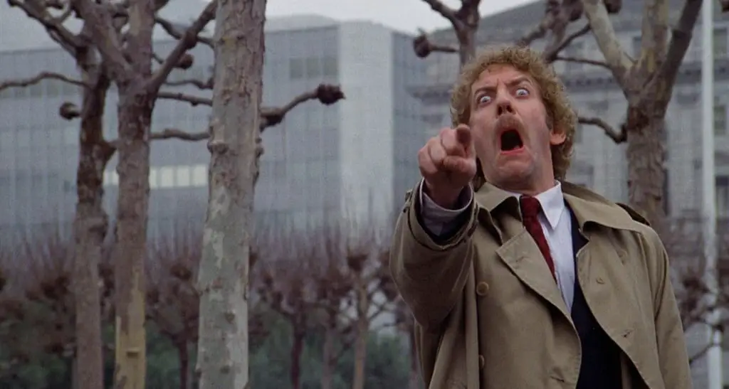 Matthew Bennell, wearing a trenchcoat in a park pointing with a look of terror on his face in Invasion of the Body Snatchers.