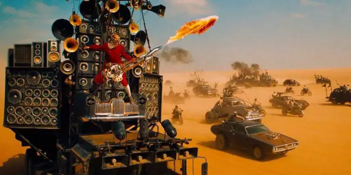 The Doof Warrior shoots flames from his guitar atop his car made of speakers. This is metal as hell.