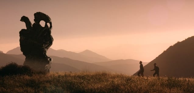 Two silhouetted figures walking a hill towards a strange rock formation