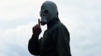 Richard wearing a gas mask with his finger to his mouth in a shushing motion