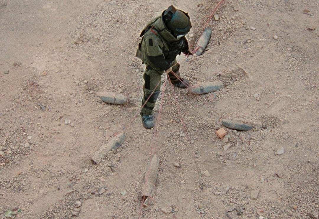 sergeant james in his bomb suit, lifting up a wire that is connected to 6 bombs that surround him