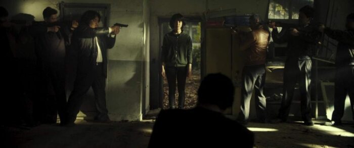 Sook-hee stands in a doorway while a half-dozen assailants aim their guns at her.