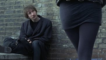 Johnny (in Mke Leigh's Naked, 1993) sits against a brick wall as unknown woman looks over at him