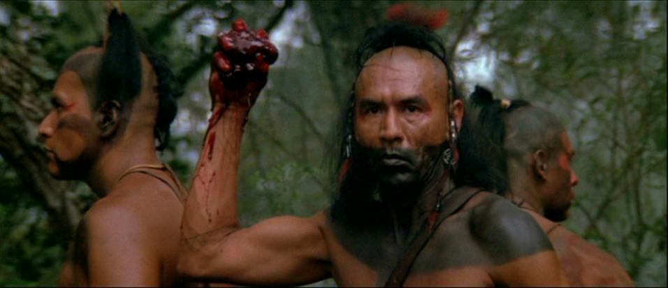 Magua holds up the heart of his enemy, his face and chest are painted in black and he is wearing a feathered head dress