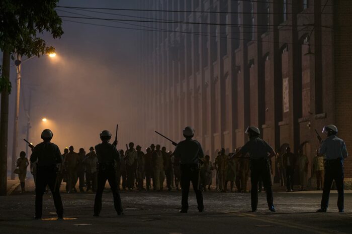 Police and rioters are seen in silhouette against the street lamps at night.
