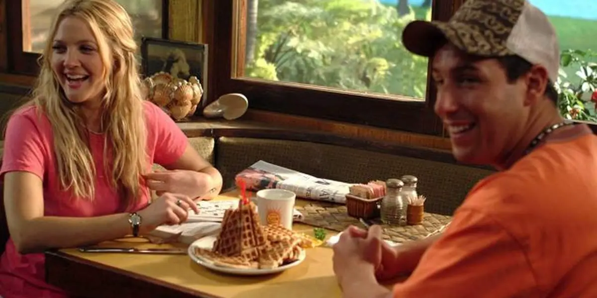 Drew Barrymore and Adam Sandler in 50 First Dates sitting at a table with a waffle tower in the center, both are looking to the side and smiling
