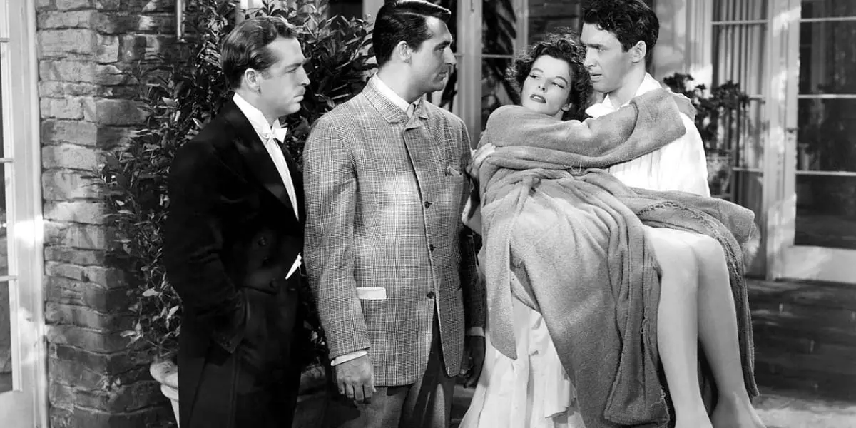 John Howard and Cary Grant stand to the left, looking at Jimmy Stewart sternly as he holds a dazed-looking Katharine Hepburn in his arms in black and white in The Philadelphia Story