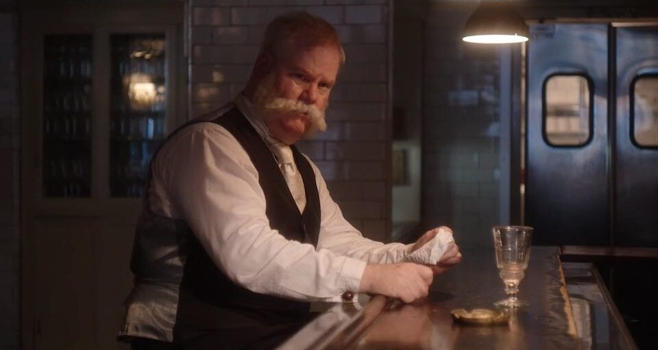 George Westinghouse (Jim Gaffigan) sits at a bar drinking a glass of lemonade. His mustache is large and white.