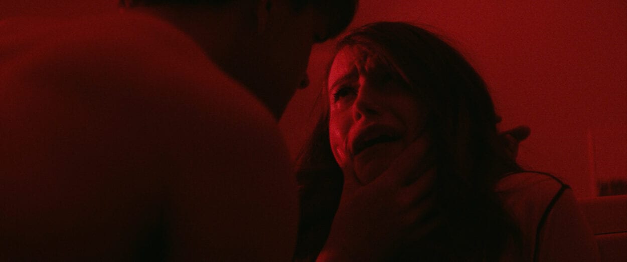 Tom (Jim Schubin) grips Eve's (Chloe Carroll) crying face in a red-lit room.